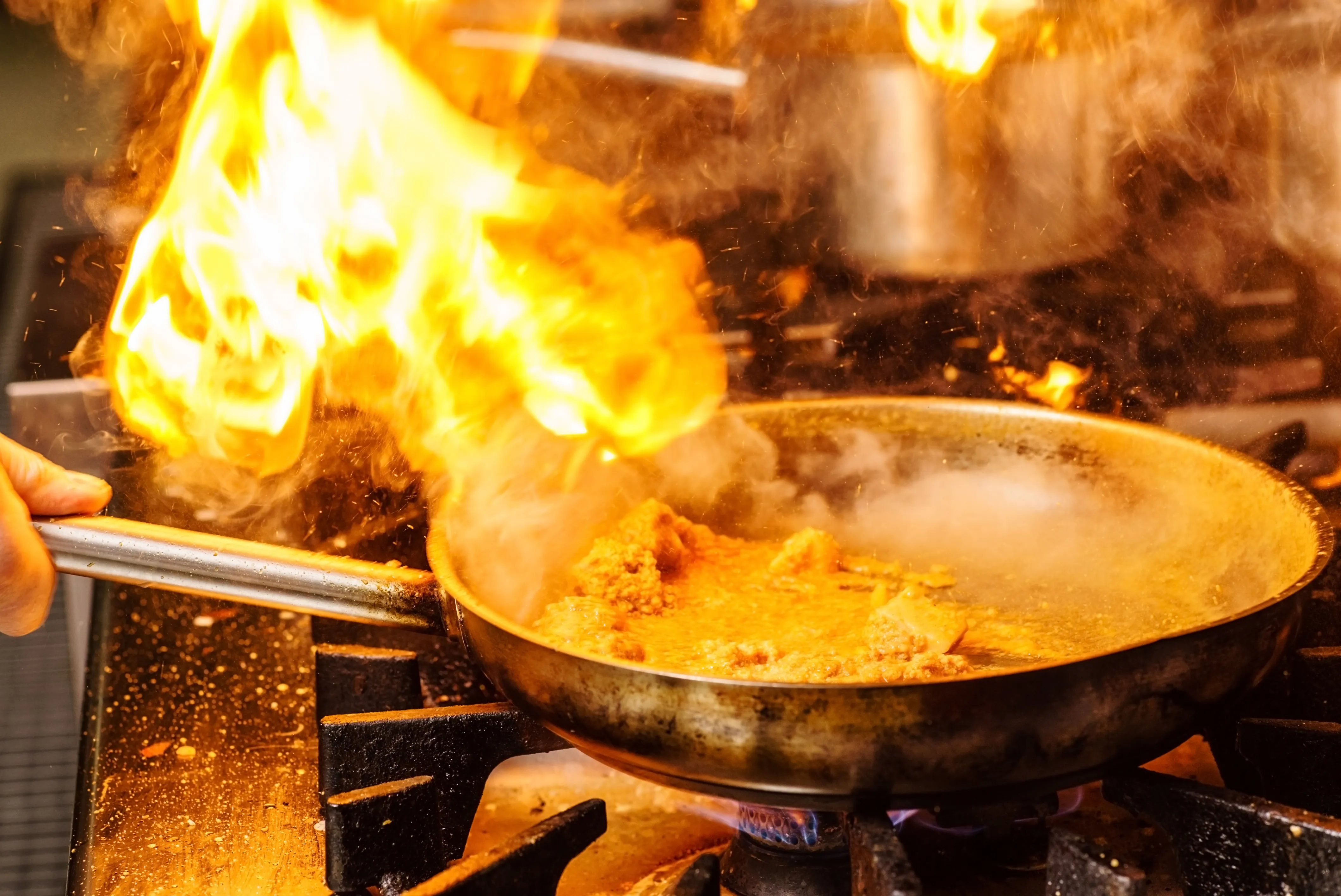 Flammen züngeln aus einer Pfanne mit kochendem Essen auf einem Gasherd, Funken und Rauch steigen auf